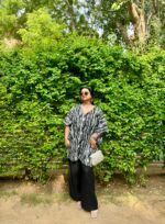 Woman wearing a black and white Shibori V-neck oversized kaftan with wide-leg black pants, standing against a vibrant green leafy backdrop.