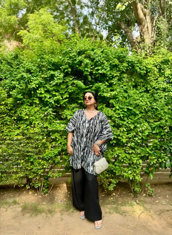 Woman wearing a black and white Shibori V-neck oversized kaftan with wide-leg black pants, standing against a vibrant green leafy backdrop.
