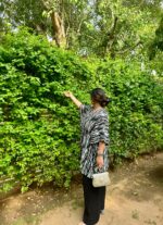 Woman wearing a black and white Shibori V-neck oversized kaftan with wide-leg black pants, standing against a vibrant green leafy backdrop.