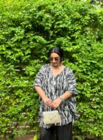 Woman wearing a black and white Shibori V-neck oversized kaftan with wide-leg black pants, standing against a vibrant green leafy backdrop.