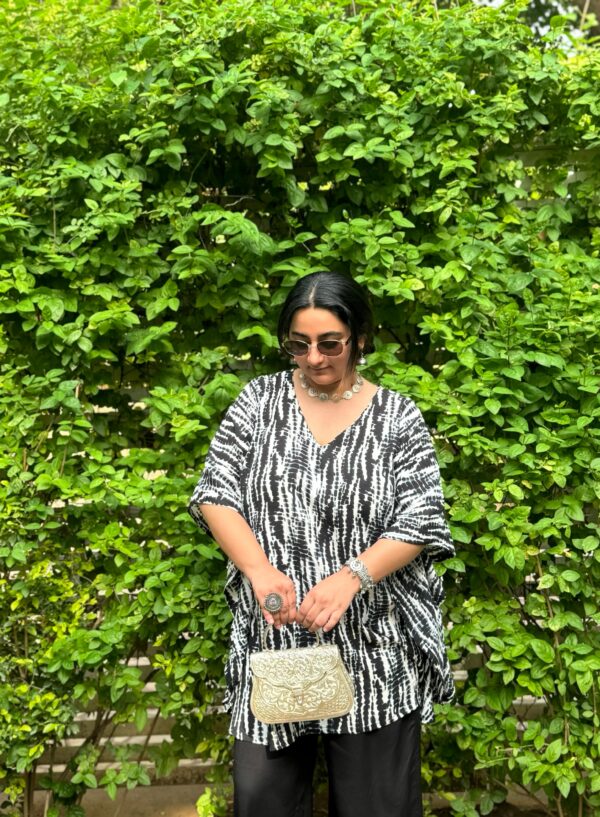Woman wearing a black and white Shibori V-neck oversized kaftan with wide-leg black pants, standing against a vibrant green leafy backdrop.