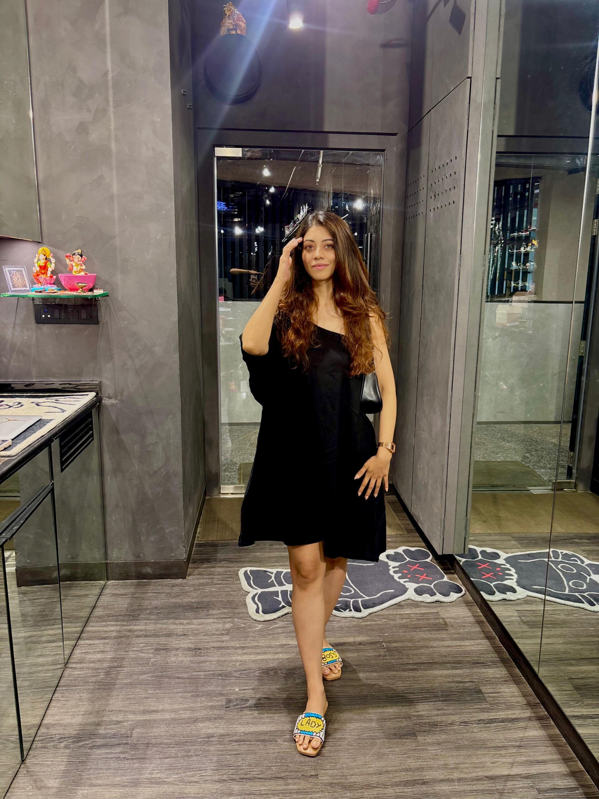 One-Shoulder Black Mini Dress Woman wearing a stylish one-shoulder black mini dress paired with colorful slip-on sandals in a modern indoor setting